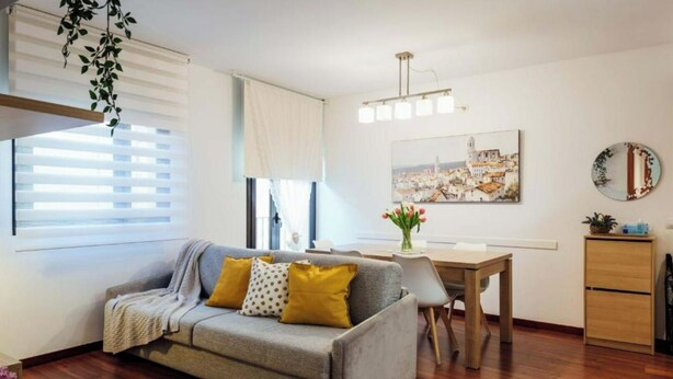 A modern living room with a grey sofa, wooden flooring, a dining table, and a painting of a cityscape. - 2 bedroom apartment RM7 (Girona)