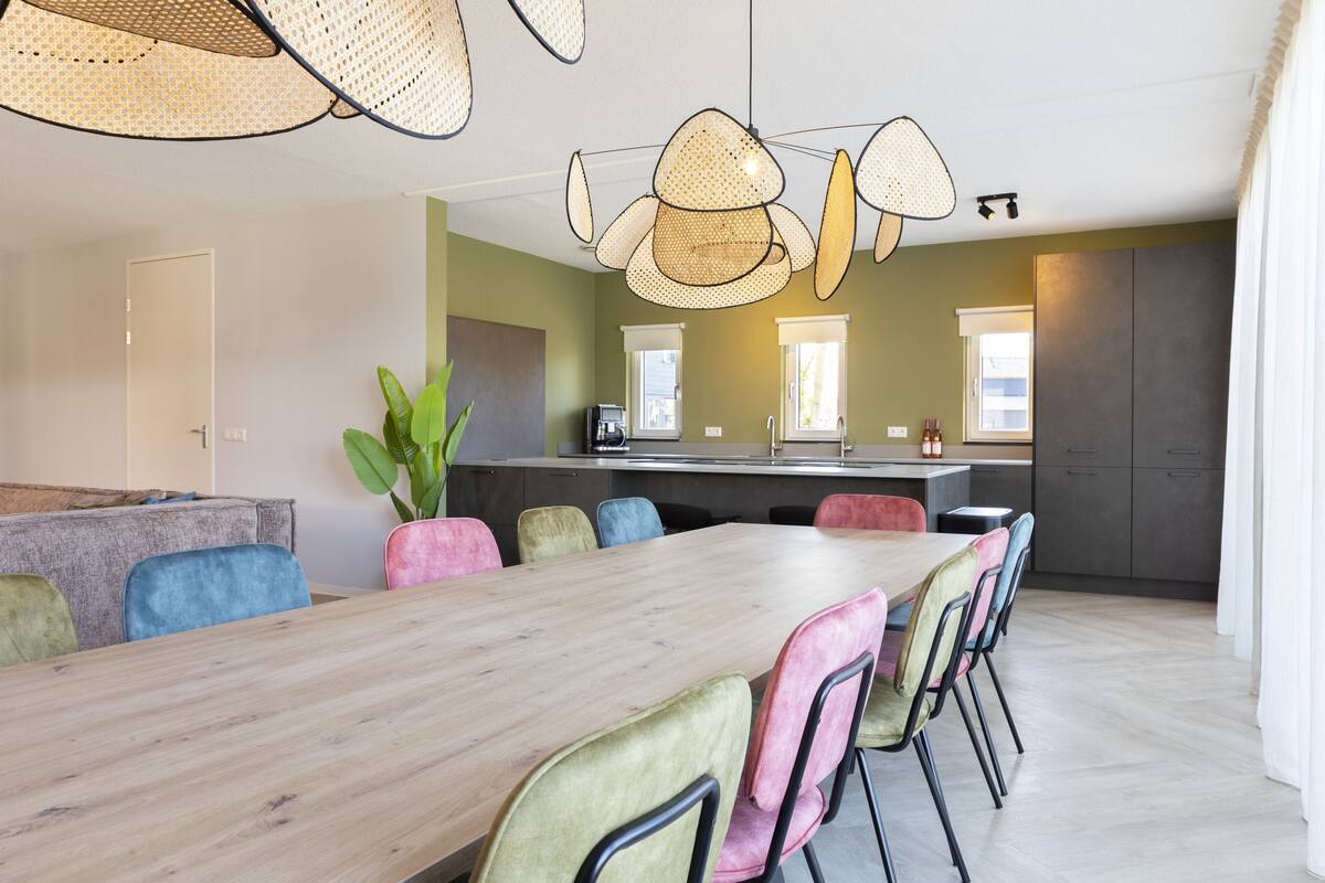 Un comedor moderno con una mesa de madera y sillas coloridas, una cocina al fondo y una planta grande.