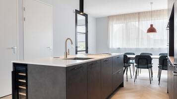 A modern kitchen with a central island, a dining area with chairs, and a large window with sheer curtains.
