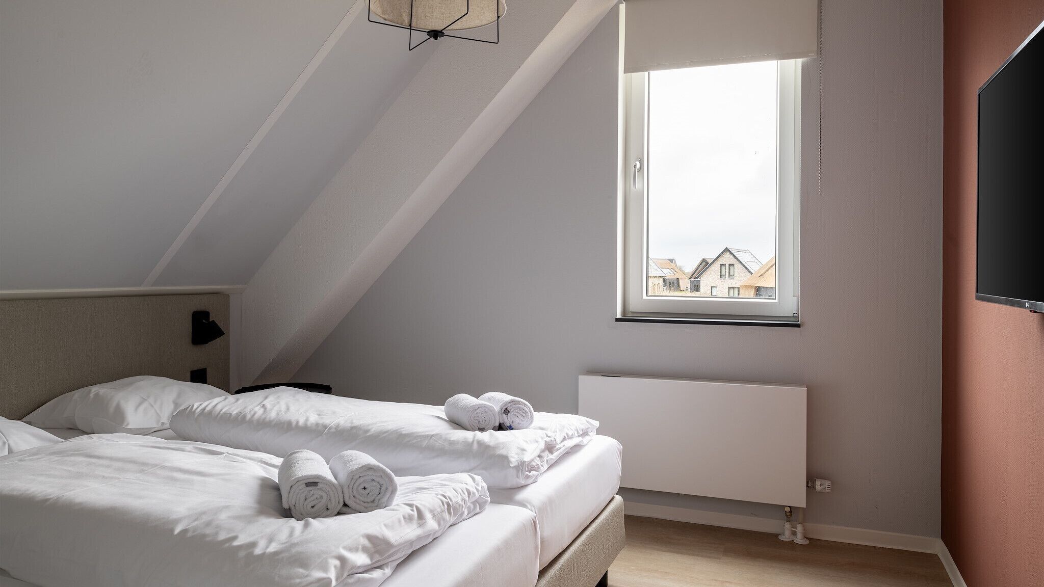 A modern, well-lit bedroom with a sloped ceiling, a large window with a view of houses, and neatly made beds with rolled towels.
