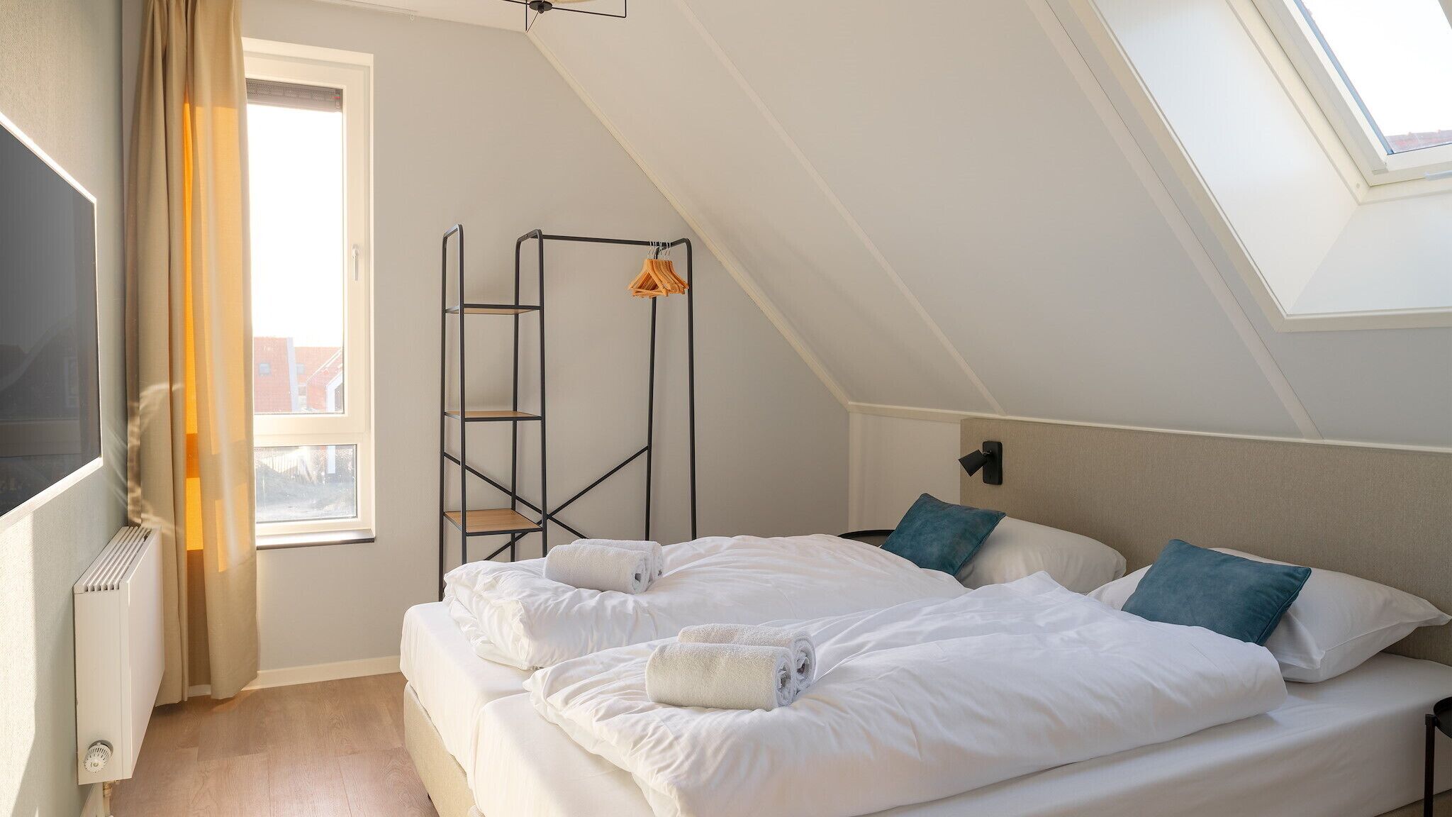 Une chambre avec un plafond en pente, un lit avec une literie blanche, deux oreillers, un radiateur et une fenêtre avec des rideaux.