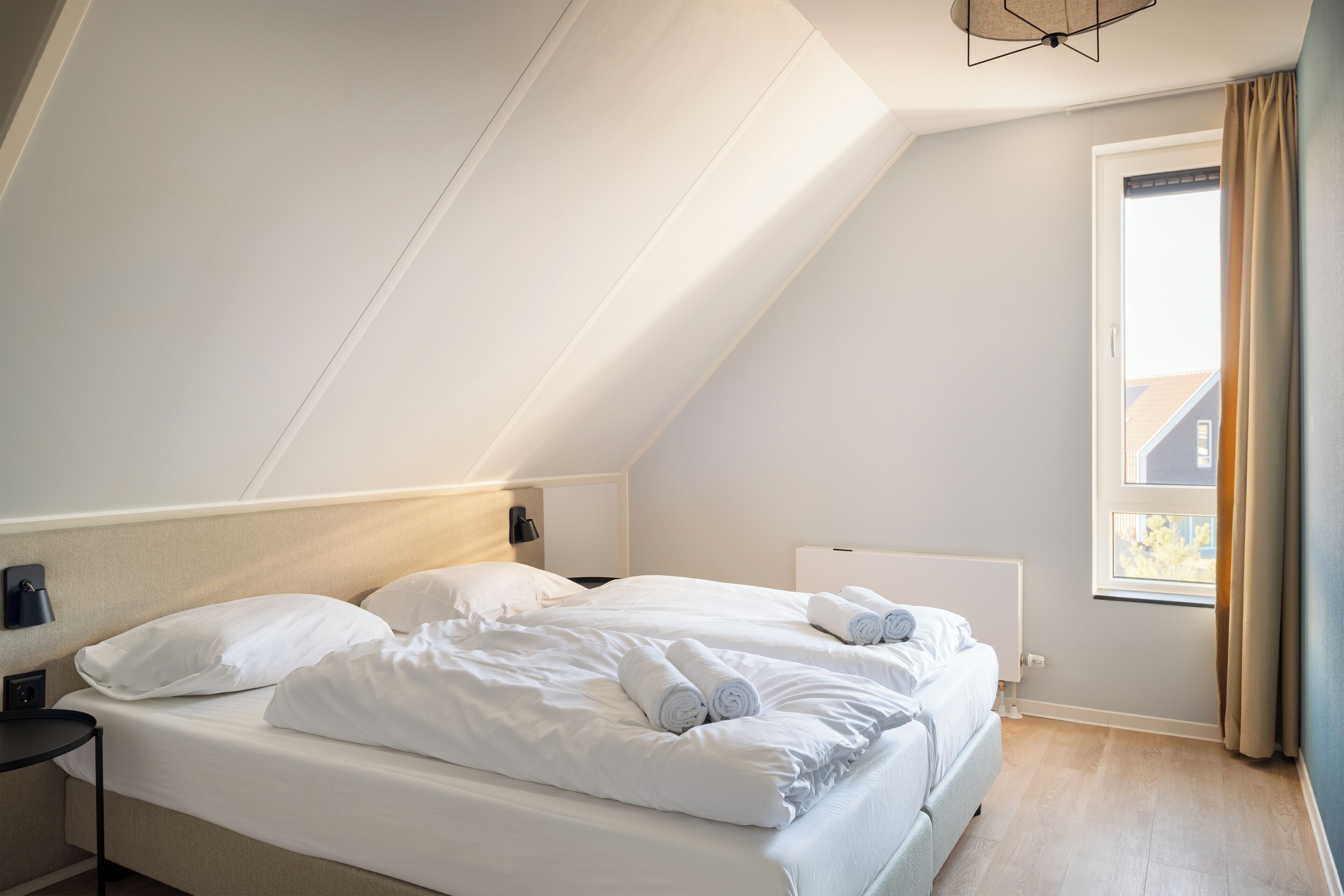 A modern, well-lit room with a large bed, a bedside table, and a window with curtains.
