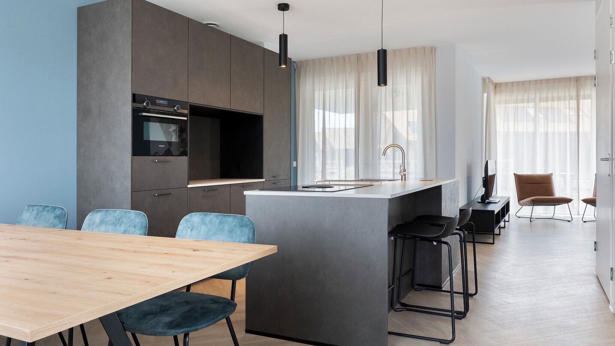 A modern kitchen with a built-in oven, a dining area with a wooden table and blue chairs, and a small island with a sink.