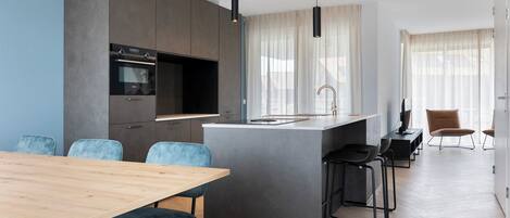 A modern kitchen with a built-in oven, a dining area with a wooden table and blue chairs, and a small island with a sink.