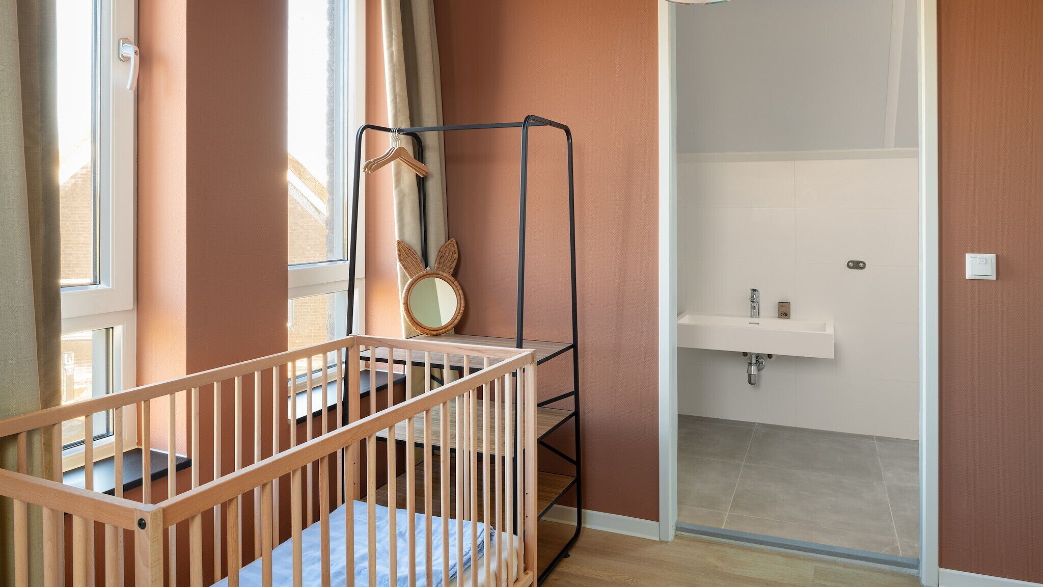 Ein Zimmer mit einem Holckrippenbett, einem Kleiderständer, einem Spiegel mit Hasenohren und einem Badezimmer mit Waschbecken und Toilette.