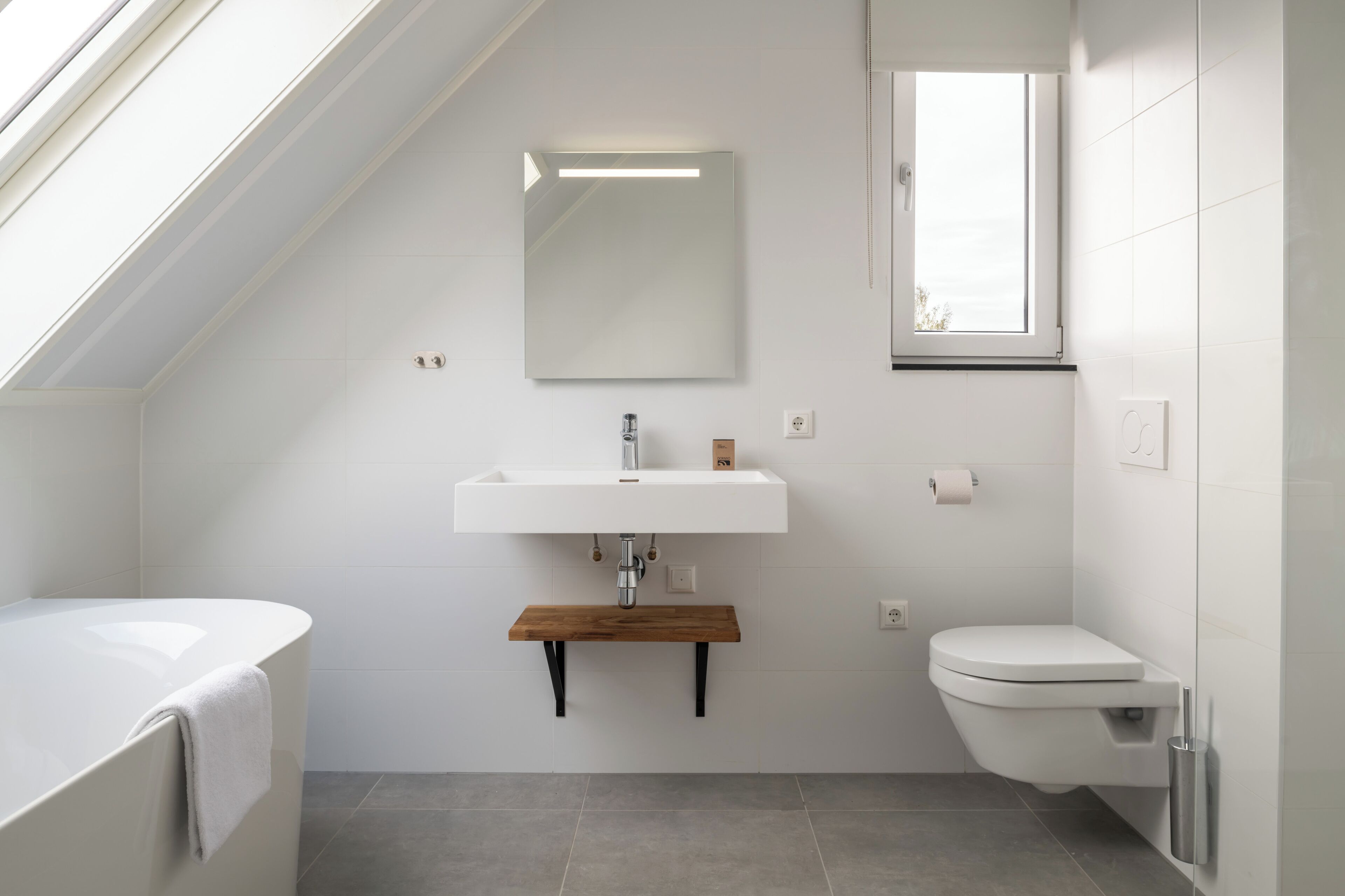 Une salle de bain moderne dotée d’une lucarne, d’une baignoire sur pieds, d’un lavabo blanc avec une étagère en bois et de toilettes.