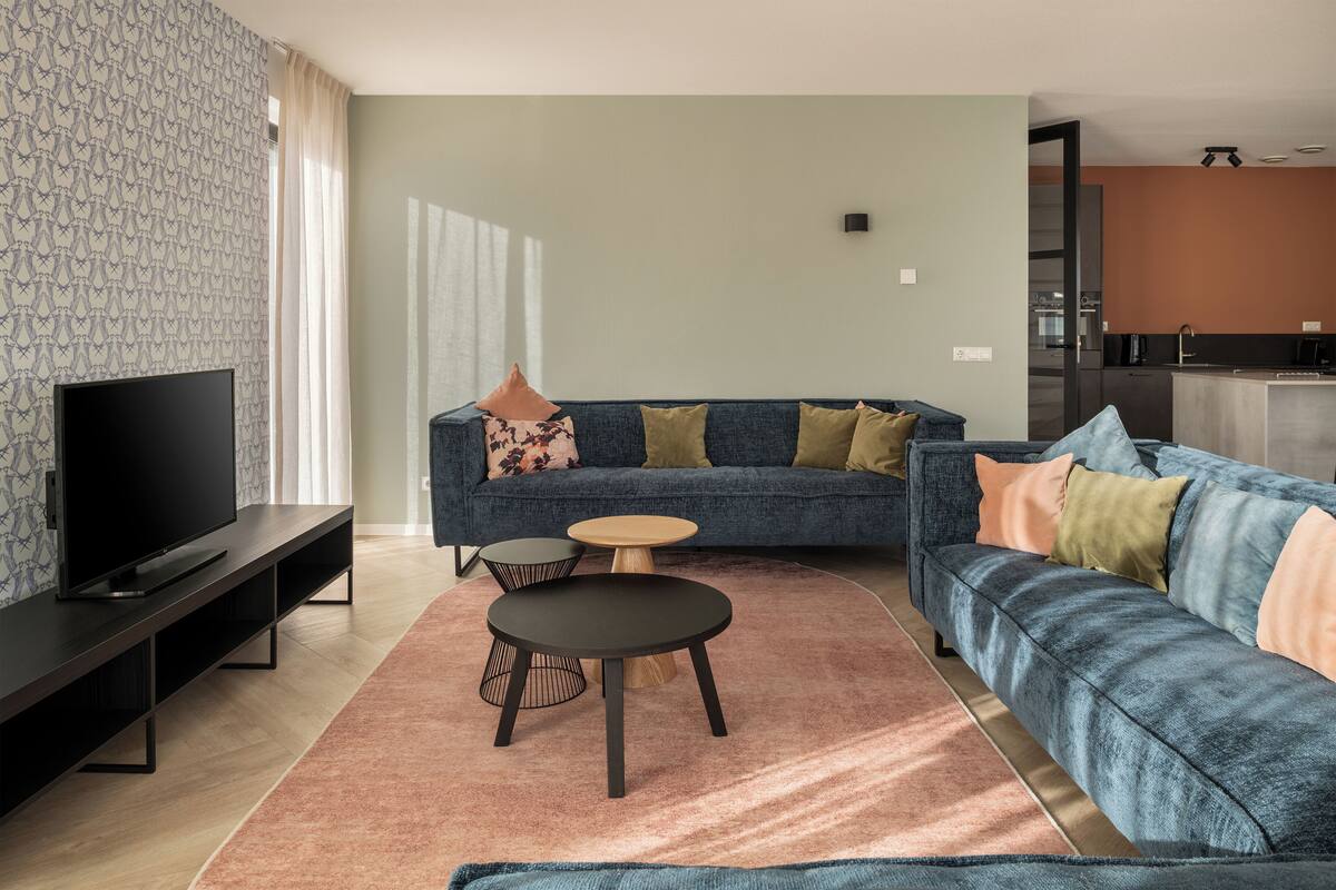 Una sala de estar moderna con un televisor de pantalla plana, un sofá azul, dos mesas redondas y una cocina al fondo.