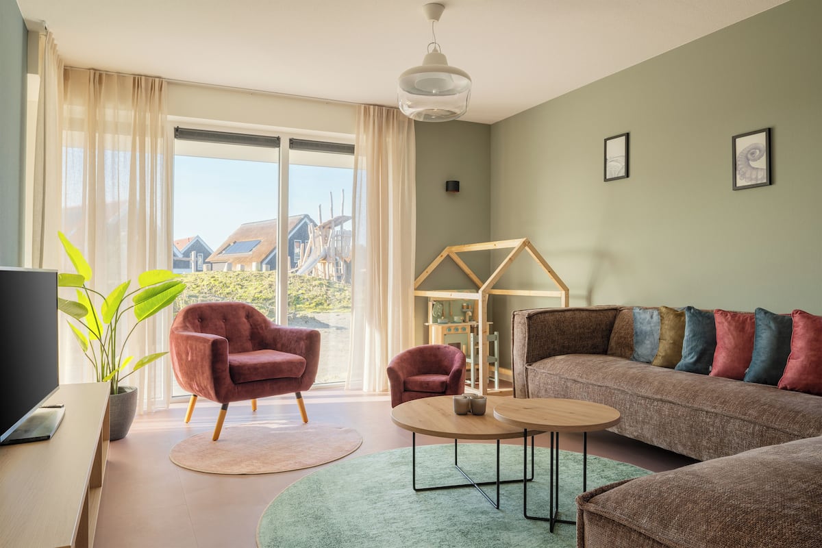 Una sala de estar con sofá, sillones, una mesa de centro y televisor. Hay una planta y una ventana con cortinas.