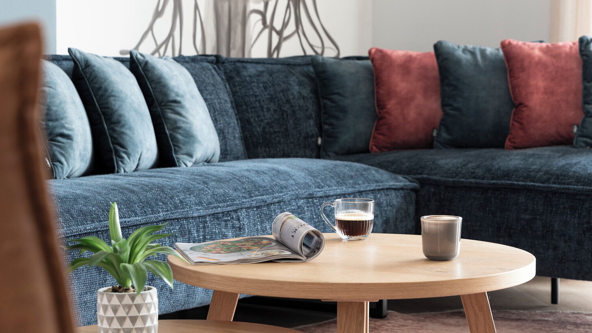 A modern living room with a blue sectional sofa, a round wooden coffee table, a magazine, a glass mug, and a potted plant.