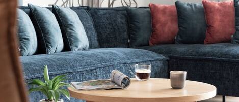 A modern living room with a blue sectional sofa, a round wooden coffee table, a magazine, a glass mug, and a potted plant.