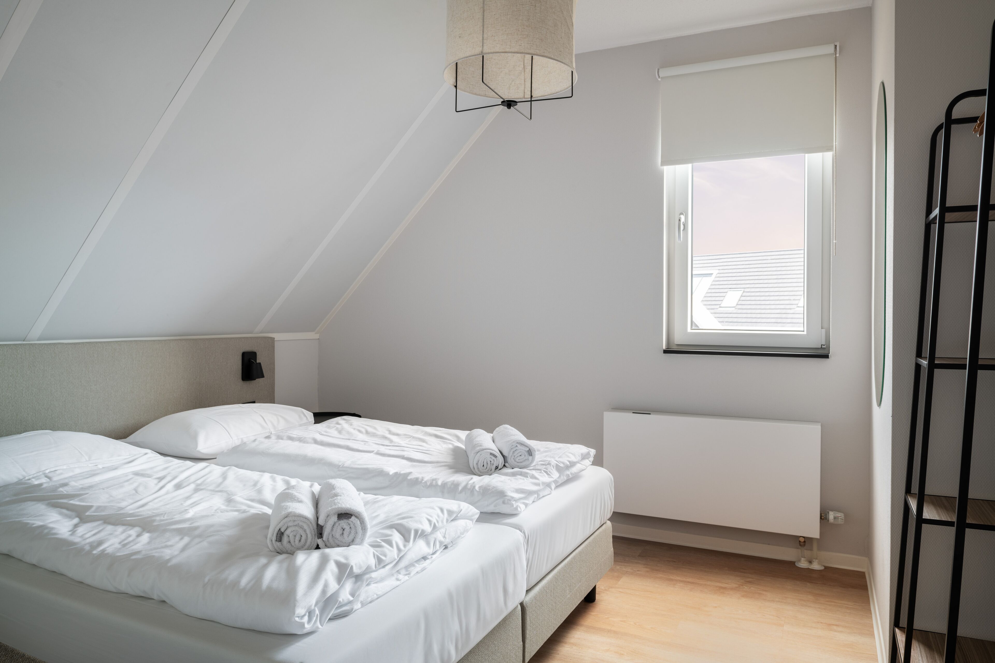 A modern, well-lit bedroom with two beds, a radiator, and a window with blinds.