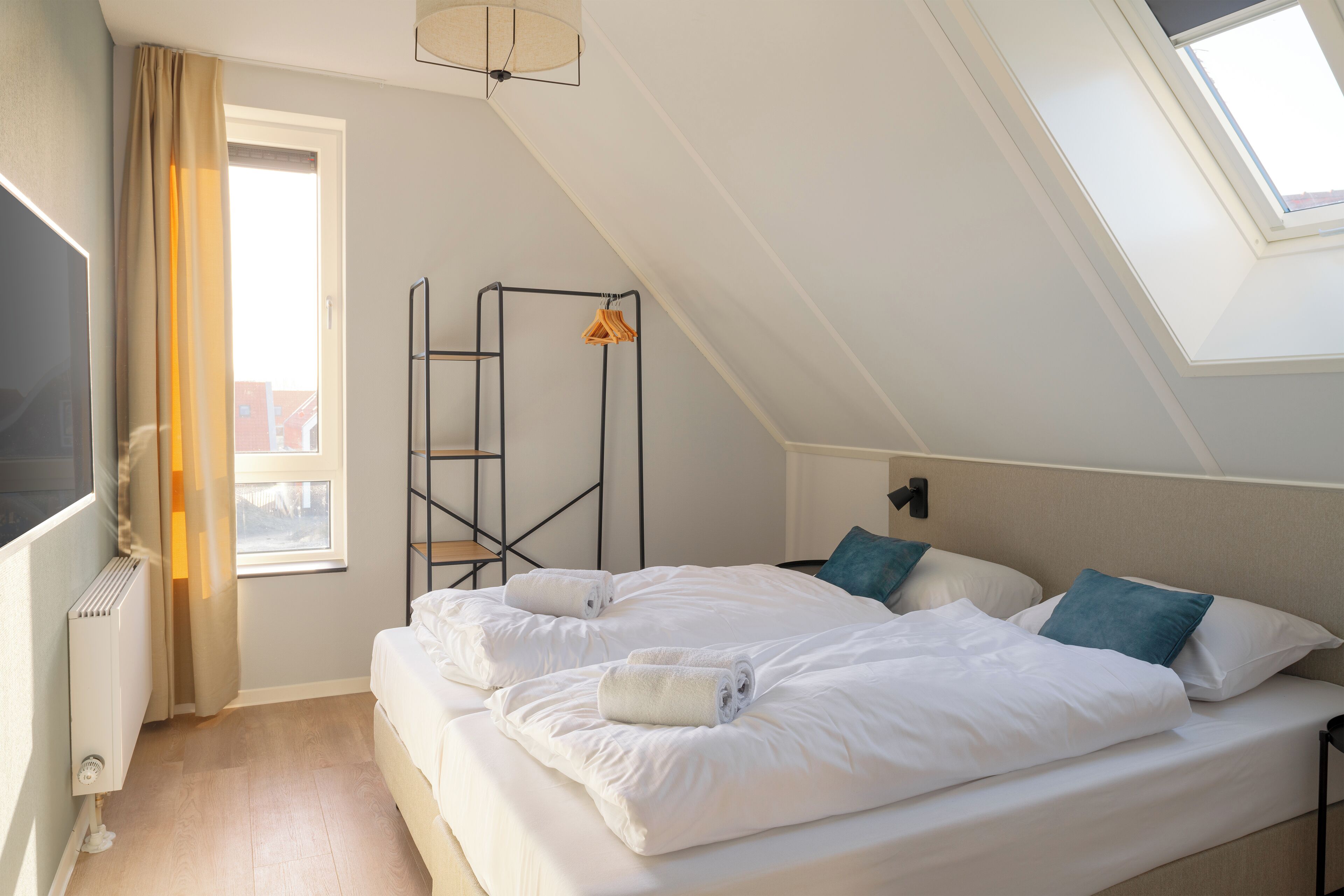 Une chambre à coucher avec un lit, deux oreillers, un radiateur, une fenêtre avec des rideaux et un puits de lumière.