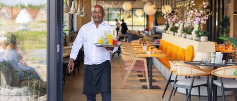 Un camarero lleva una bandeja con bebidas y una fuente de frutas, caminando por un restaurante con suelo de madera, mesas redondas y sillas.