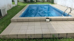 A rectangular swimming pool with a tiled edge, surrounded by a grassy area and a white wall. - Detached house with garden and indoor parking (Abidjan)