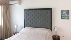 A bedroom with a tufted headboard, a bed with a white bedspread, two bedside tables with lamps, an air conditioning unit, and a painting on the wall. - Detached house with garden and indoor parking (Abidjan)