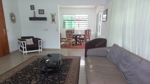 A living room with a beige sofa, a white armchair, a glass coffee table, and a patterned rug. There is a dining area in the background with a table and chairs. - Detached house with garden and indoor parking (Abidjan)