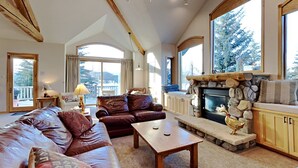 A cozy living room with a stone fireplace, leather sofas, and large windows offering a view of the outdoors. - Beautiful home with stunning views, a hot tub, and a pool table ready! (Dillon)