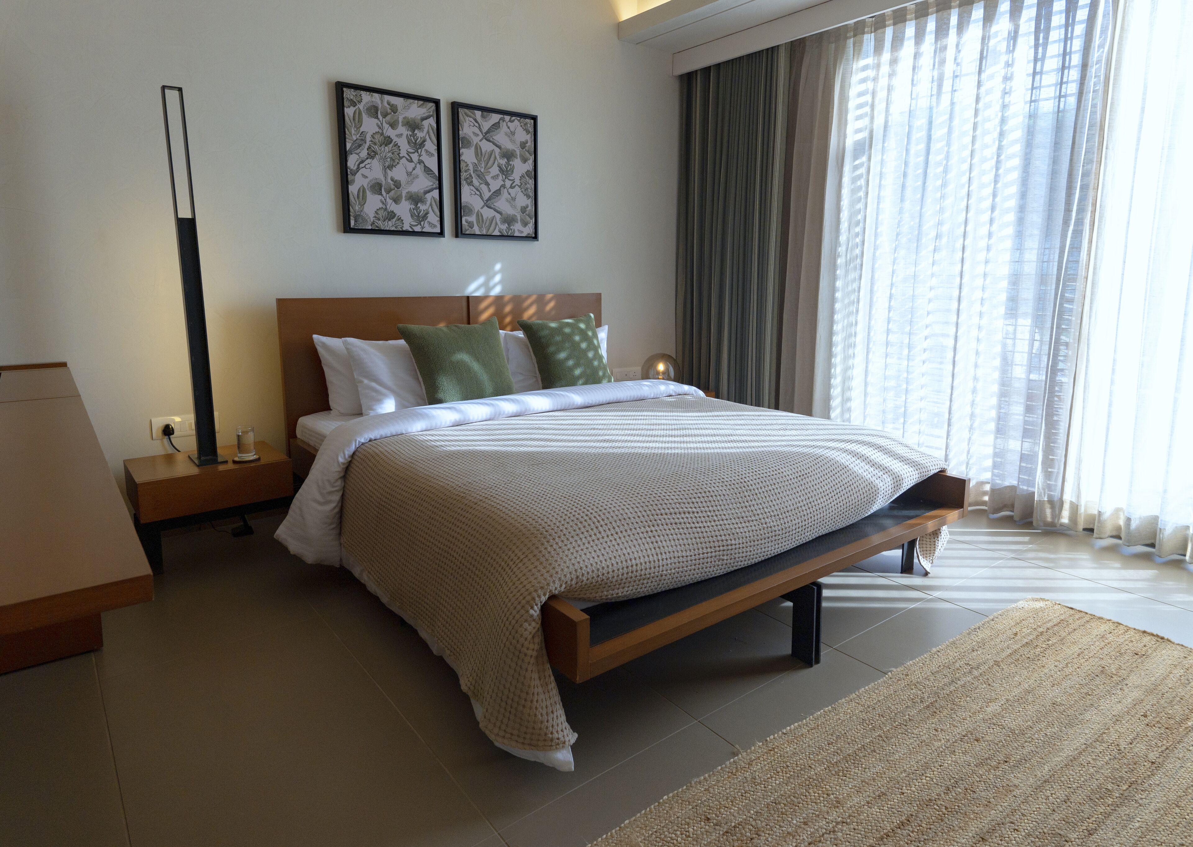 A modern bedroom with a large bed, a nightstand, a bedside table, a lamp, and two framed botanical prints on the wall.