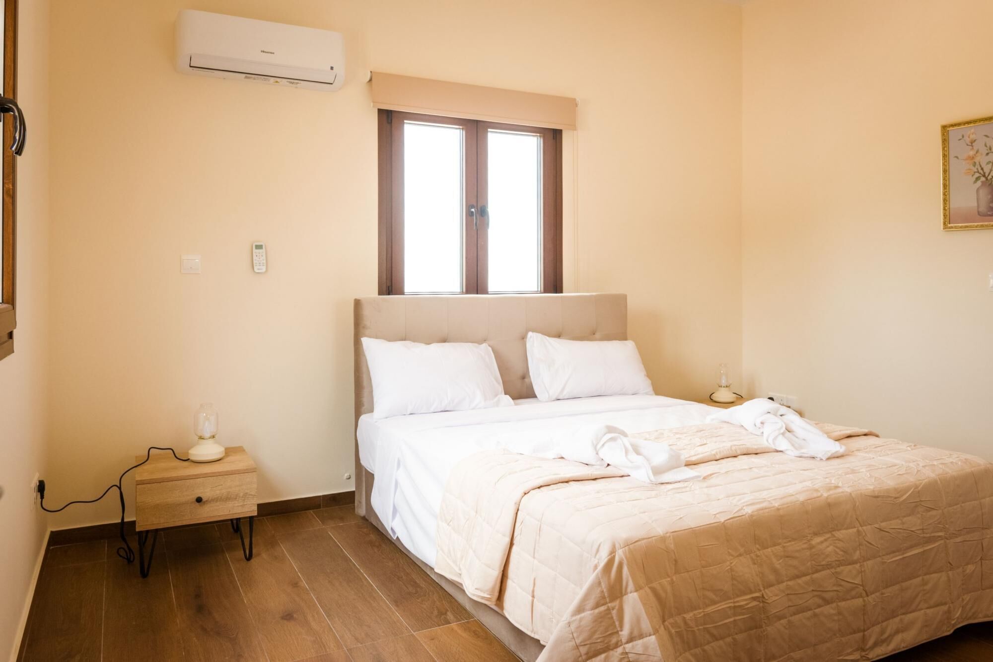 Un dormitorio con cama, mesitas de noche, aire acondicionado, una ventana y un cuadro en la pared.