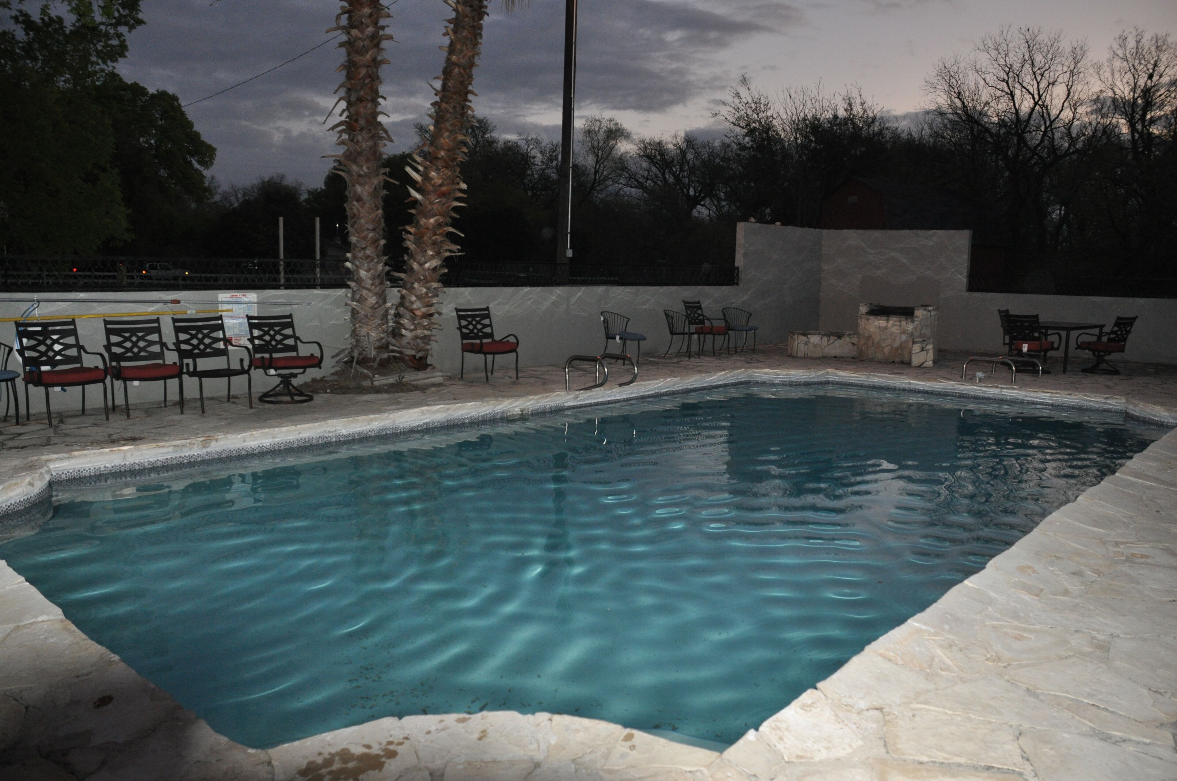 A rectangular swimming pool with clear water, surrounded by stone tiles and equipped with multiple chairs and a table.