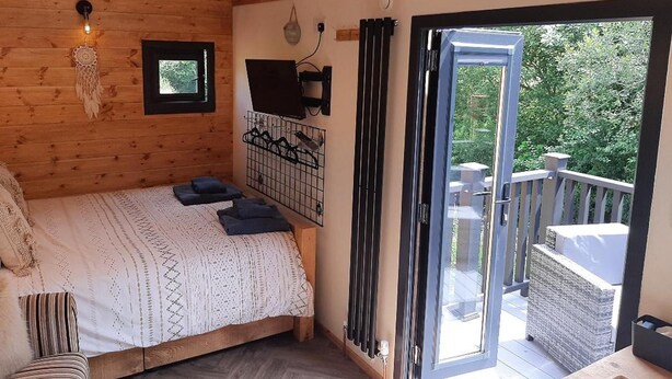 A wooden cabin room with a bed, a small window, a wall-mounted TV, and a sliding glass door leading to an outdoor balcony.
