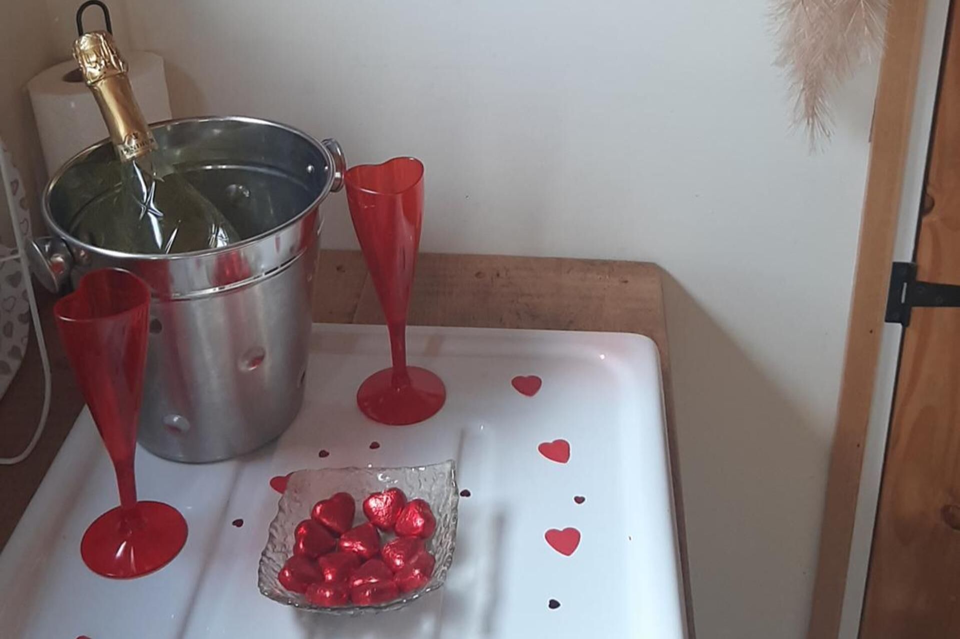 A table with a metal bucket, two red glasses, and a glass bowl with heart-shaped chocolates.
