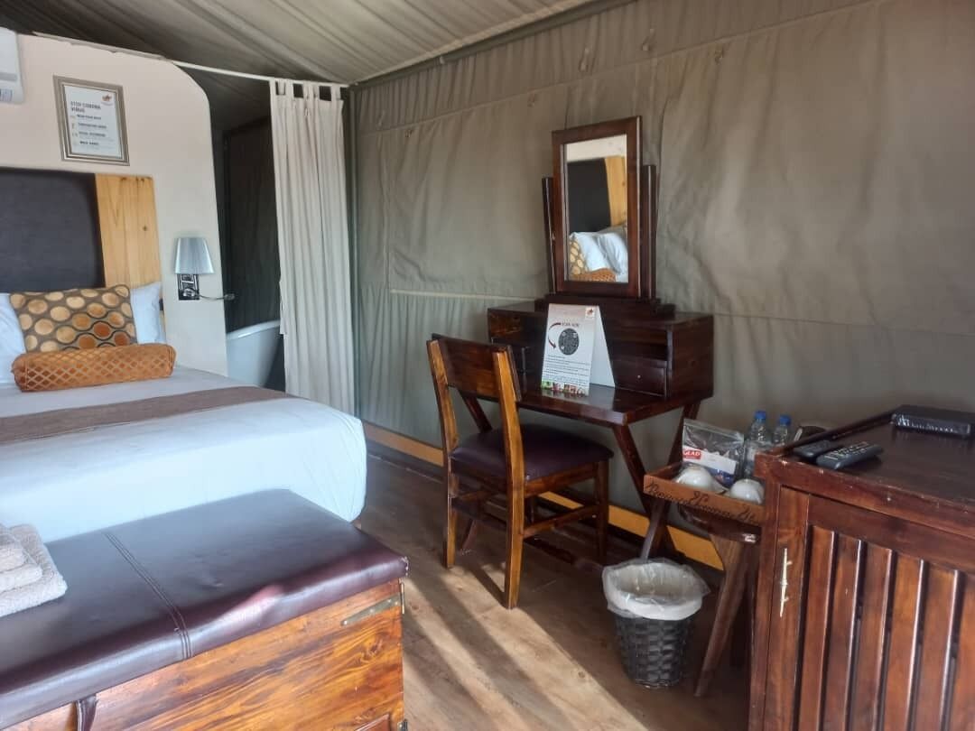 Quarto de hotel com uma cama, uma escrivaninha de madeira, uma cadeira, um cesto de lixo pequeno e um espelho.