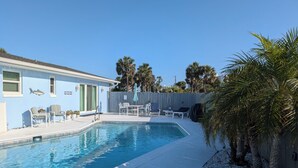 A swimming pool with lounge chairs and a table, surrounded by a fence and palm trees. - Steps from the beach with pool! (Ormond Beach)