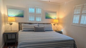 A bedroom with a bed, two nightstands, two wall-mounted lamps, and two paintings. - Steps from the beach with pool! (Ormond Beach)