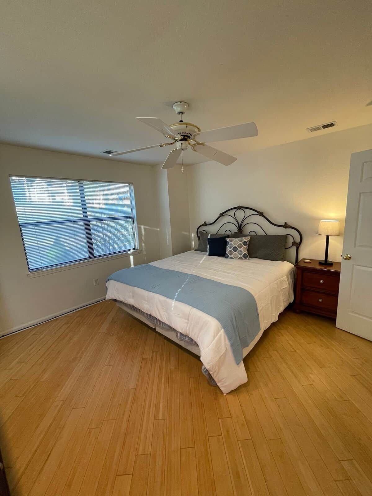A bedroom with a bed, a ceiling fan, a window with blinds, a nightstand, and a door.