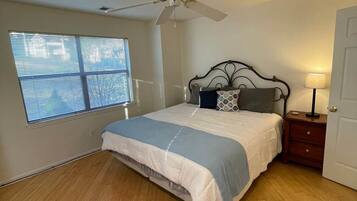 A bedroom with a bed, a ceiling fan, a window with blinds, a nightstand, and a door.
