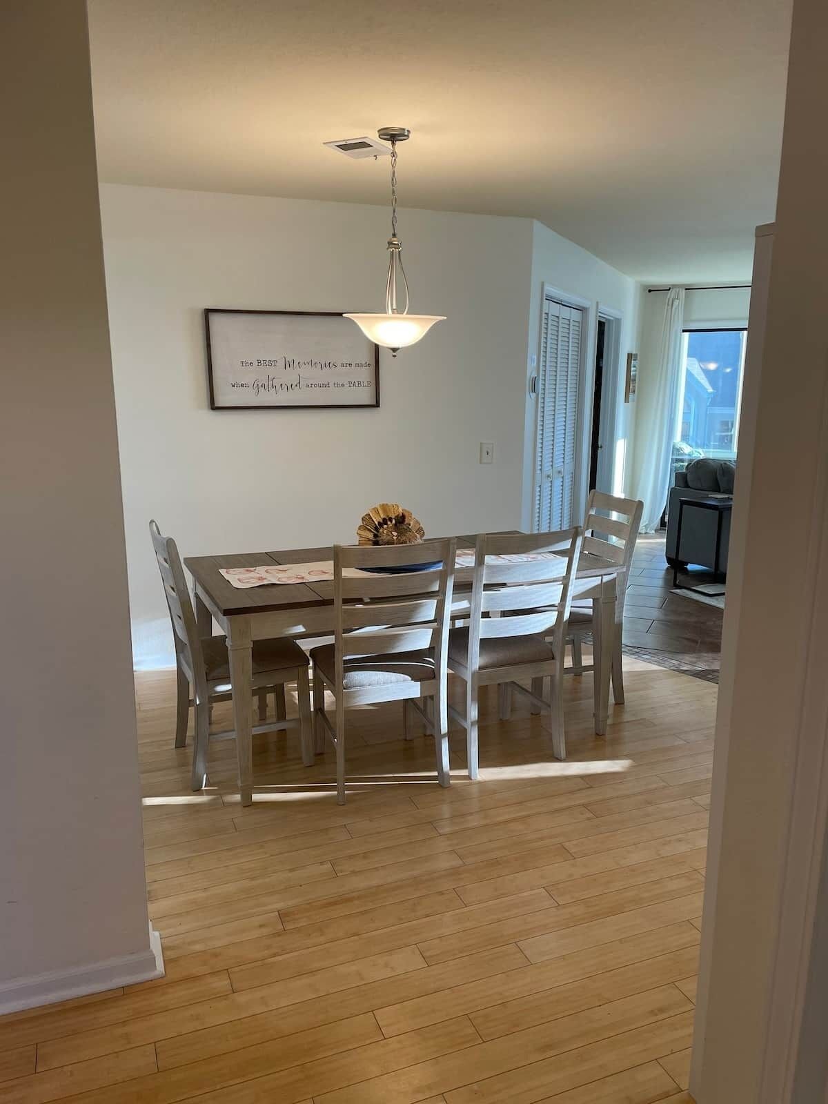 A dining area with a wooden table and chairs, a pendant light, and a framed quote on the wall.