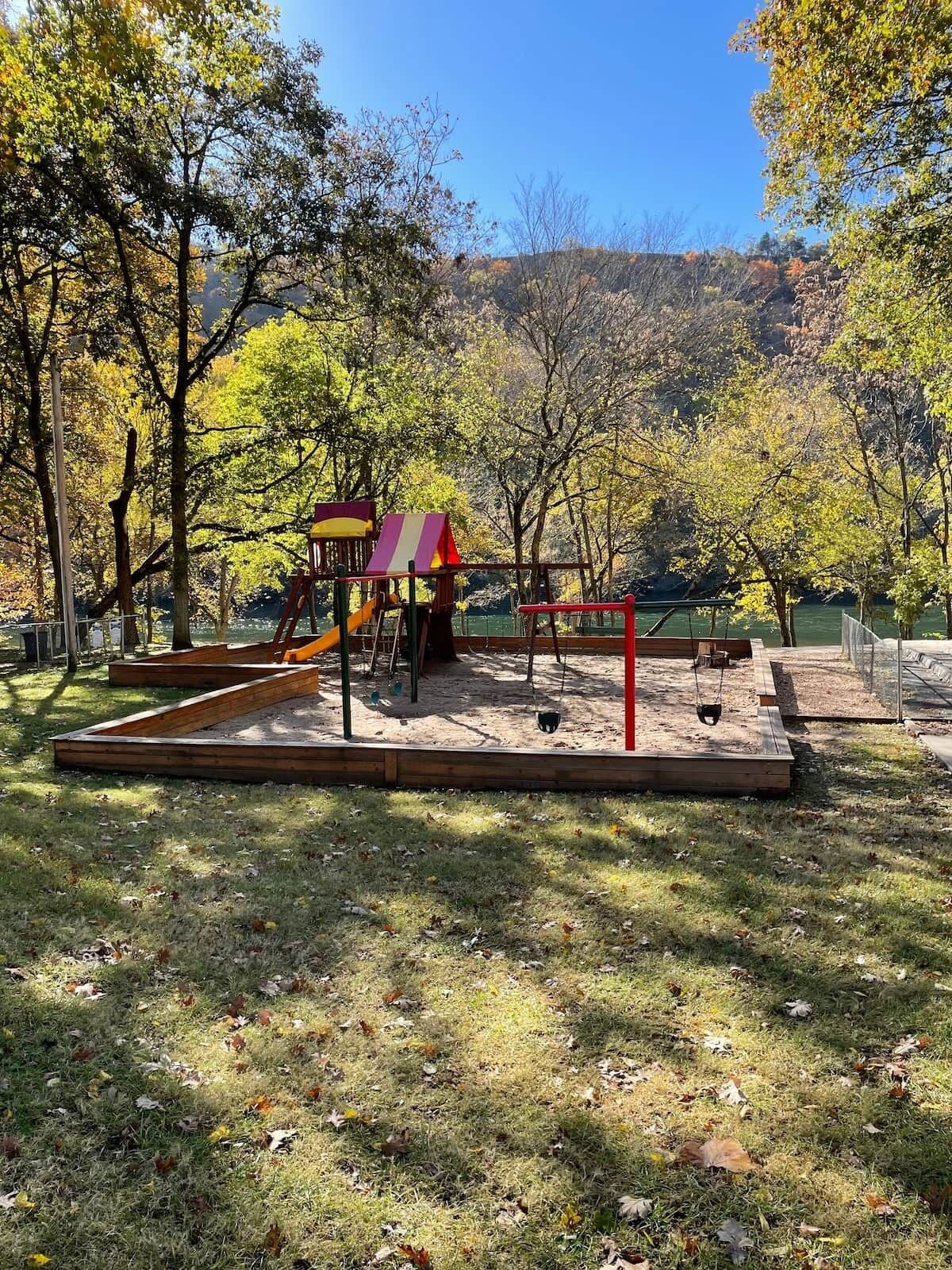 A playground with a slide and swings, surrounded by trees and a hilly landscape.