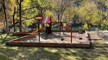 A playground with a slide and swings, surrounded by trees and a hilly landscape.