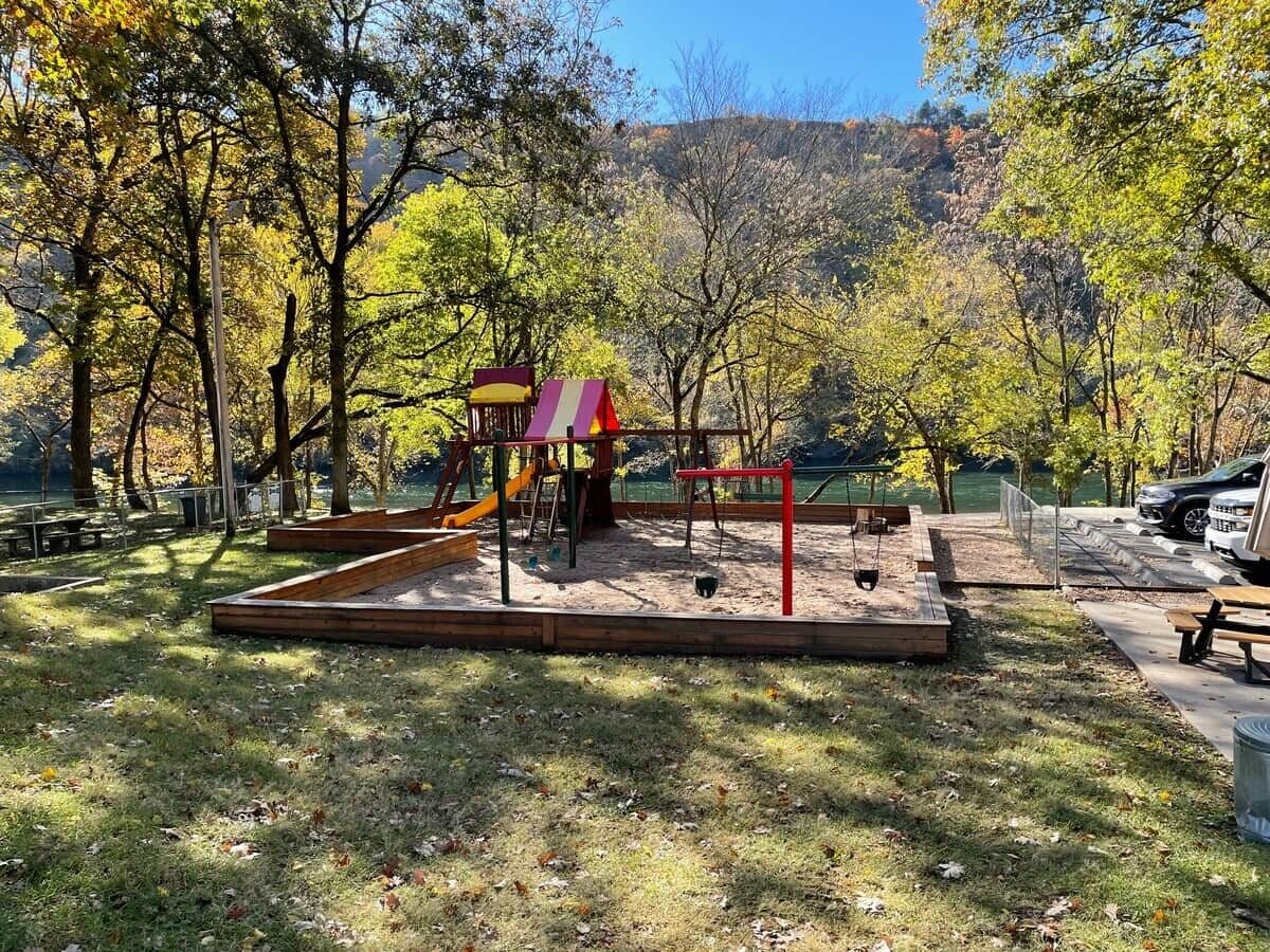 Une aire de jeux avec un toboggan et des structures d’escalade, entourée d’arbres et comprenant un espace pique-nique.