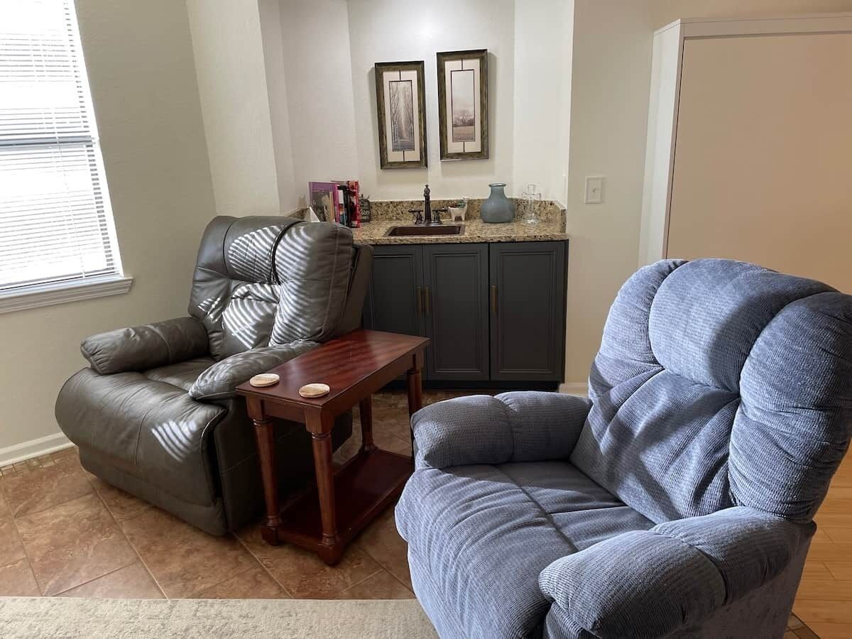 A living room with a leather recliner, a blue armchair, a wooden coffee table, and a kitchen area with a sink and framed artwork.