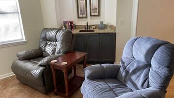 A living room with a leather recliner, a blue armchair, a wooden coffee table, and a kitchen area with a sink and framed artwork.