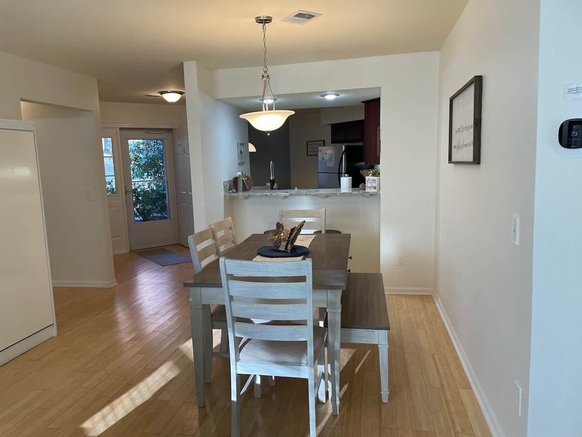 A dining area with a wooden table and chairs, a hanging light fixture, and a view into the kitchen.
