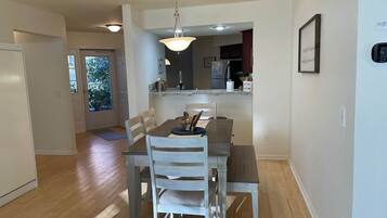 A dining area with a wooden table and chairs, a hanging light fixture, and a view into the kitchen.