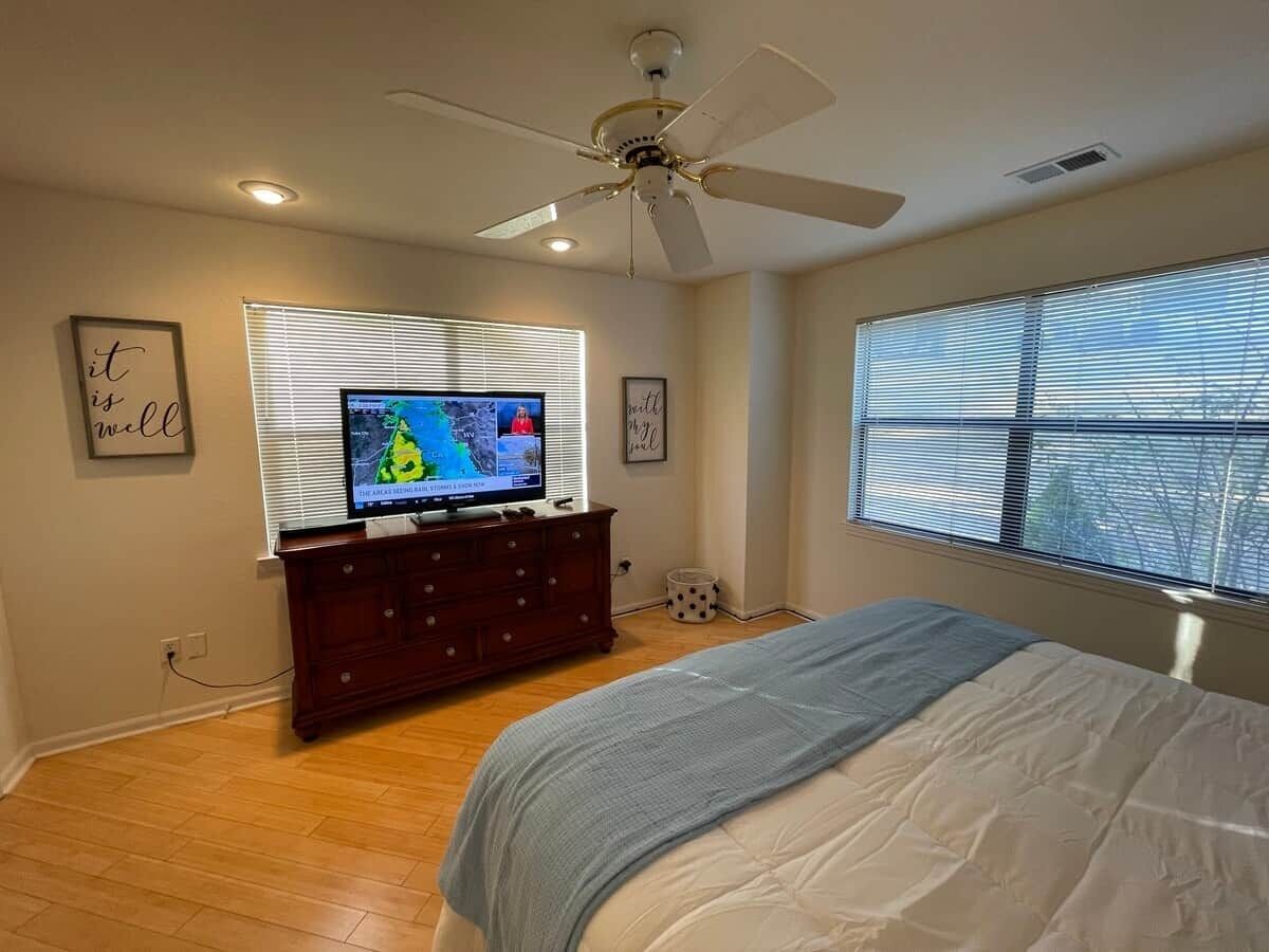 A bedroom with a bed, a dresser, a television on a stand, a ceiling fan, and a window with blinds.