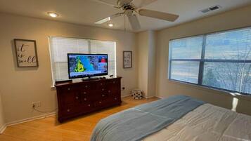 A bedroom with a bed, a dresser, a television on a stand, a ceiling fan, and a window with blinds.