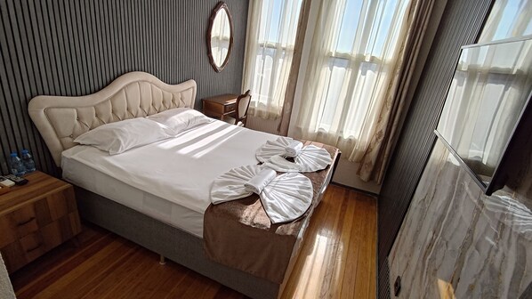 A neatly made bed with white linens and two towels folded at the foot, a wooden nightstand with a remote control and water bottles, a wooden chair, a mirror on the wall, and a window with sheer curtains. - Nuray Hotel (Istanbul)