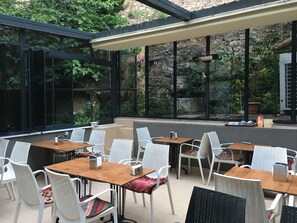 An outdoor dining area with white wicker chairs and wooden tables, surrounded by large windows offering views of greenery and a stone wall. - Nuray Hotel (Istanbul)