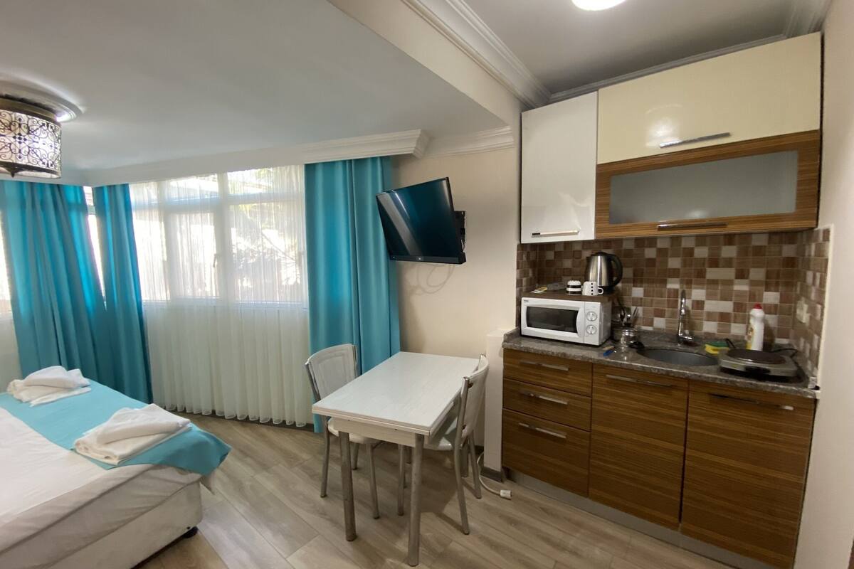 A compact hotel room with a bed, a small dining table, a TV mounted on the wall, and a kitchenette with a microwave and a coffee maker.