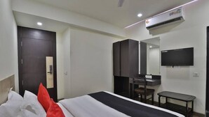 A hotel room with a bed, a desk, a chair, a TV, and an air conditioning unit. - Hotel Grand Ambience (Gandhidham)