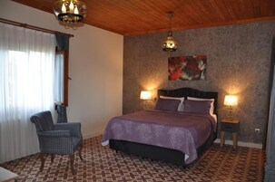 Un dormitorio con alfombra estampada, una silla a cuadros, una cama con colcha morada, dos lámparas de noche y un cuadro en la pared.