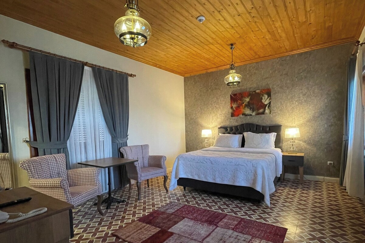 A hotel room with a wooden ceiling, a bed with white linens, a patterned rug, a checkered chair, a small table, and two wall-mounted lamps.