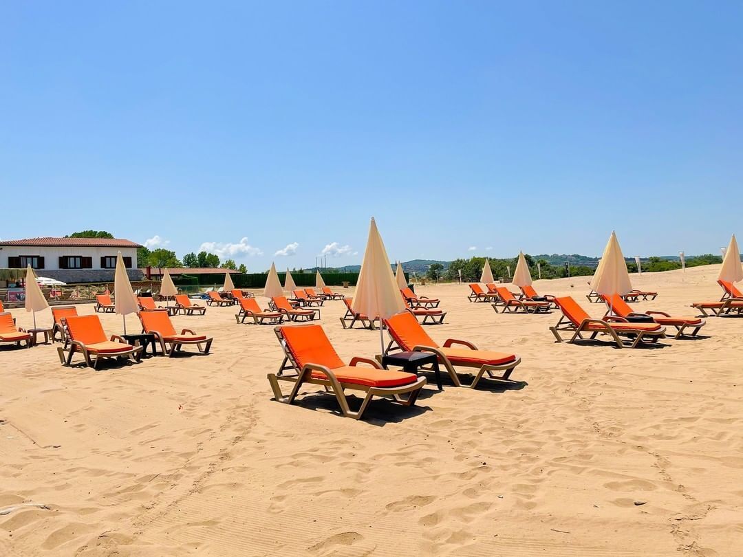 A beach with rows of orange lounge chairs and beige umbrellas, a building in the background, and a clear blue sky.