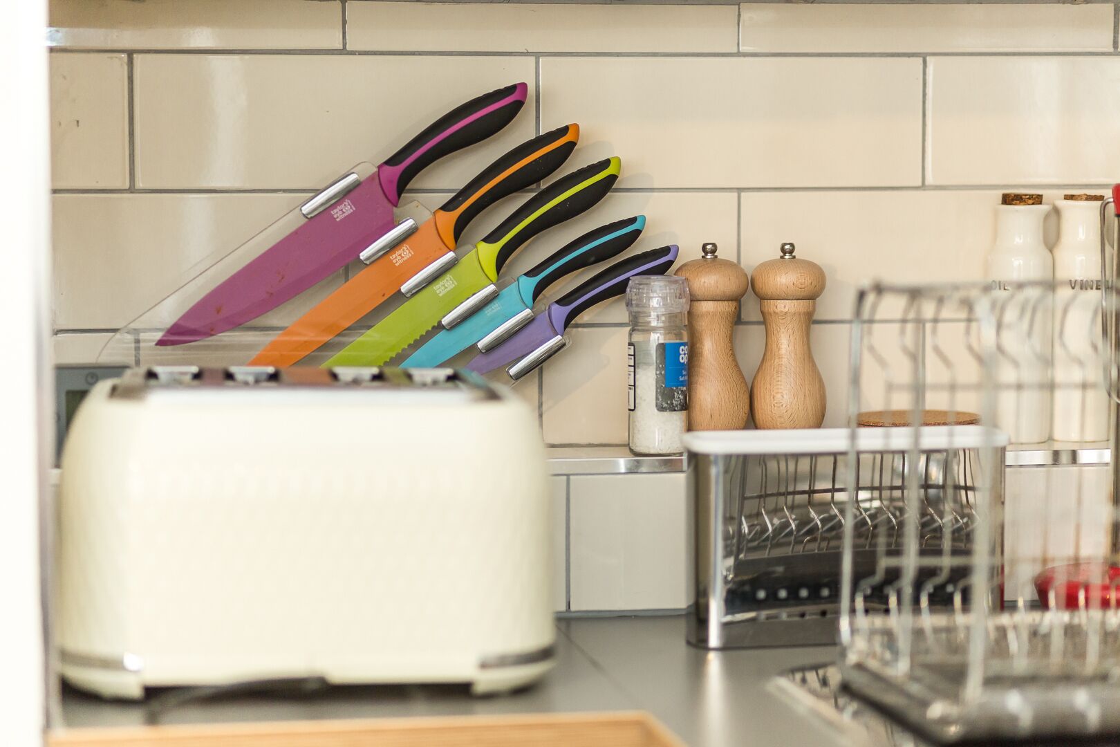 A kitchen with a white toaster, a dish rack, and a variety of colorful knives on a magnetic strip.