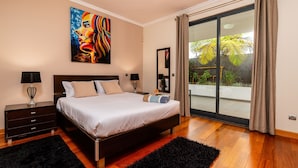 A bedroom with a large bed, a nightstand, a lamp, a painting, and a sliding glass door to a balcony with plants.